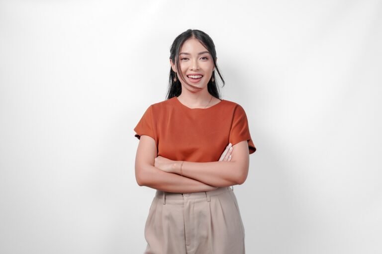 Confident young Asian woman in brown shirt and taupe pants is smiling and crossing her arms
